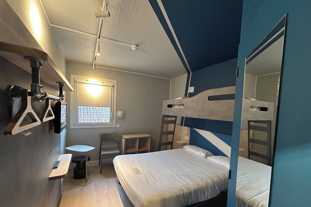 A compact hotel room with bunk beds, a small desk, and a window with blinds.