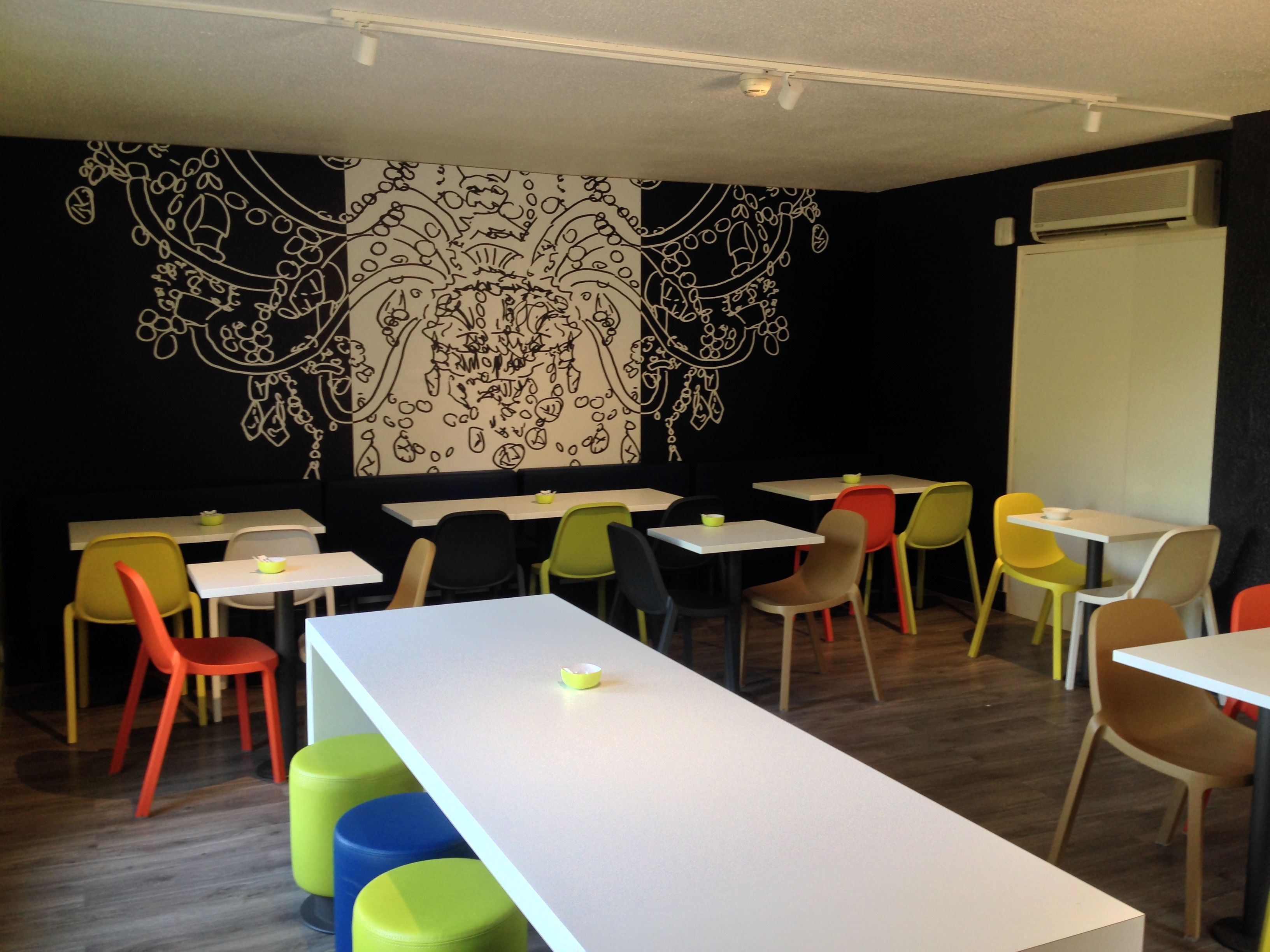 A modern dining area with white tables and colorful chairs, a black and white mural on the wall, and an air conditioning unit on the ceiling.