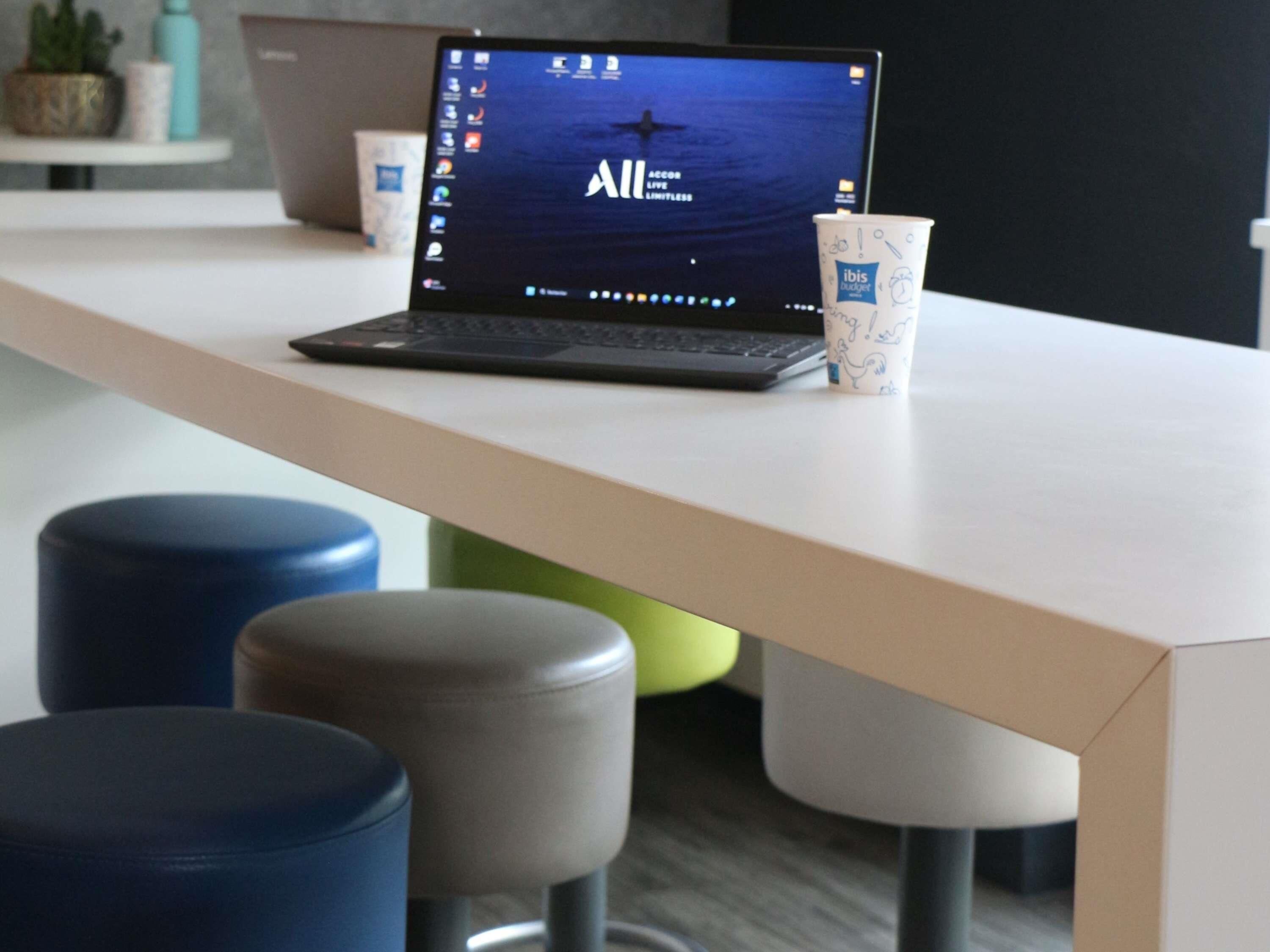 A laptop on a white table with a cup and stools in the foreground.