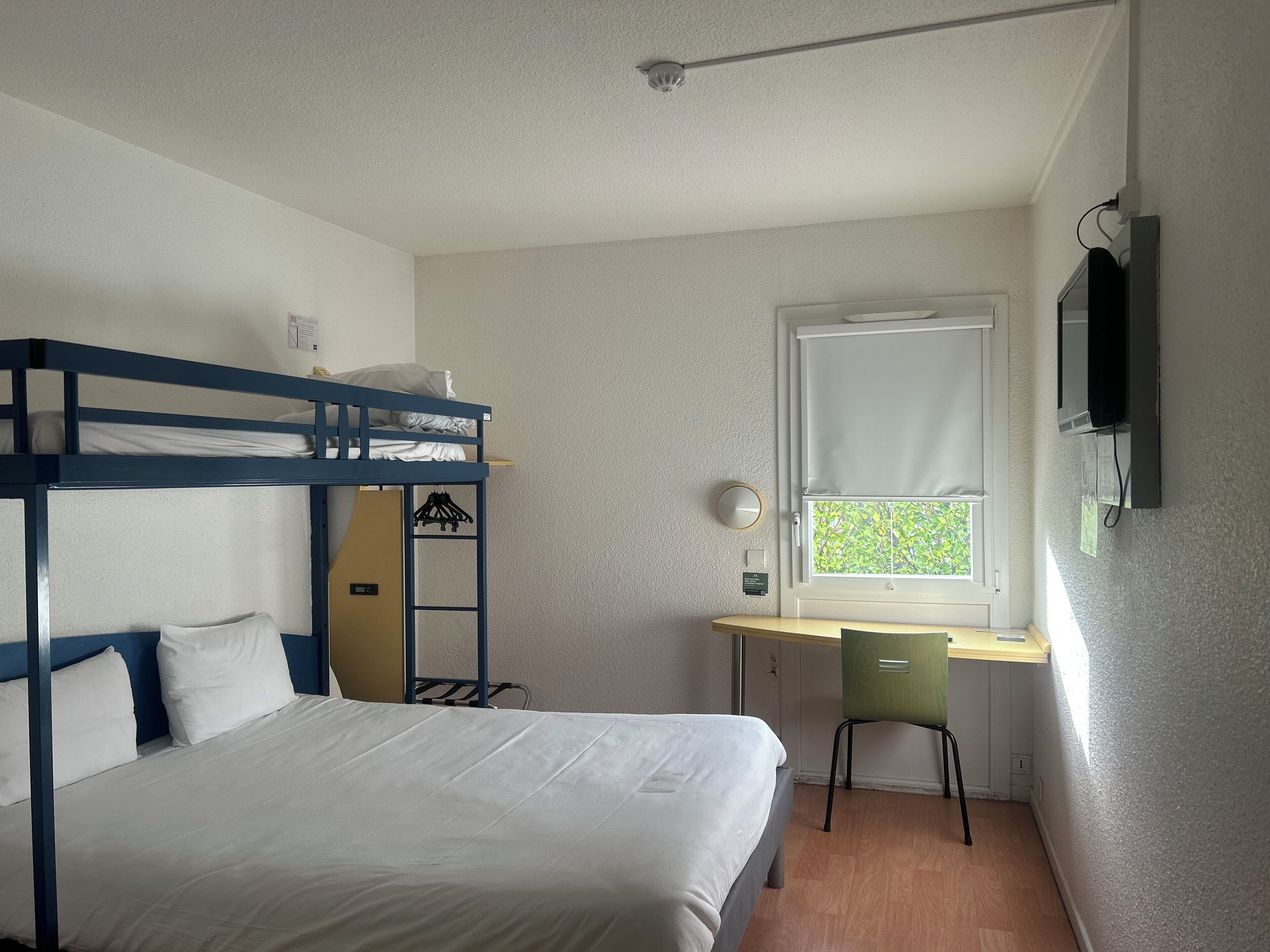 Una cama litera con ropa de cama blanca, un escritorio con una silla, una ventana con persianas y un televisor montado.