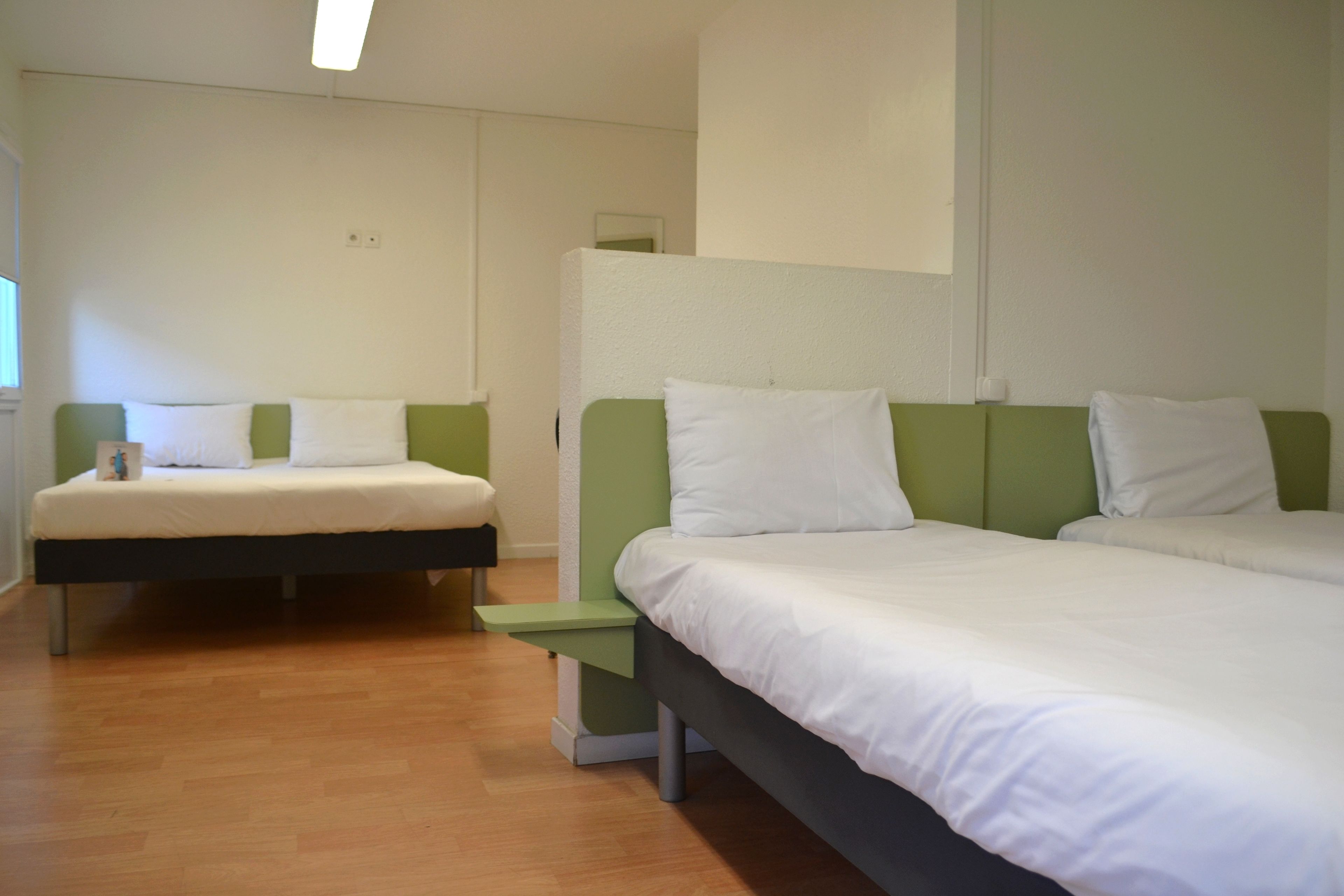 Habitación de hotel con dos camas, cada una con ropa blanca y cabeceros verdes. El piso es de madera y las paredes son blancas.