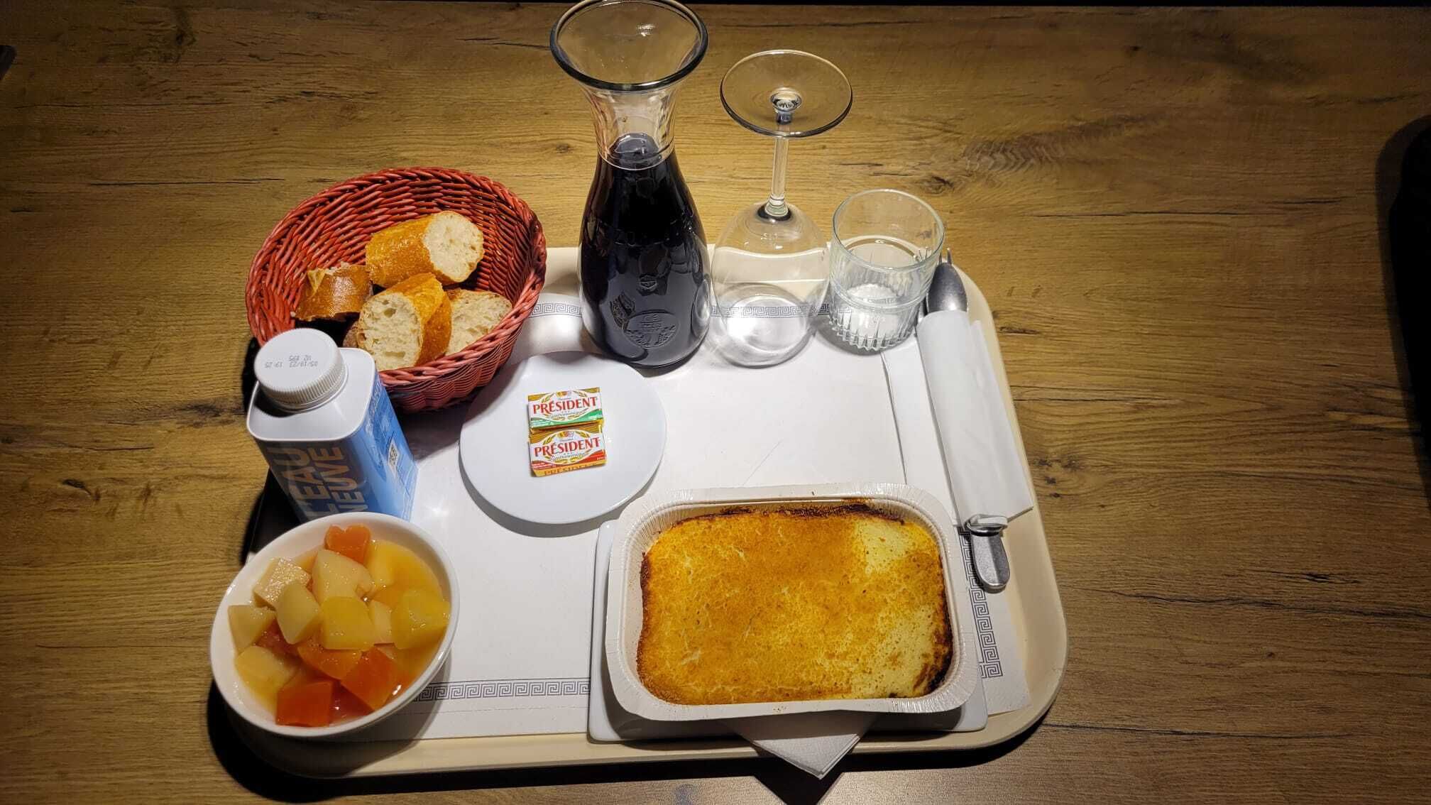 Una bandeja con un cesto de pan, una botella de agua, un vaso de agua, una copa de vino, una botella de jugo, un cuenco de ensalada de frutas y un plato al horno.
