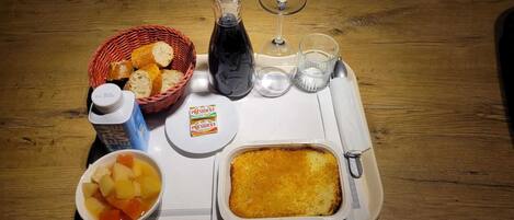 Una bandeja con un cesto de pan, una botella de agua, un vaso de agua, una copa de vino, una botella de jugo, un cuenco de ensalada de frutas y un plato al horno.