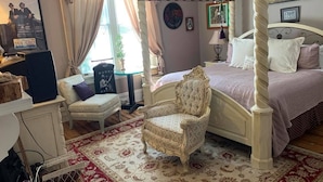 A bedroom with a four-poster bed, a chair, a desk, and a window with curtains.