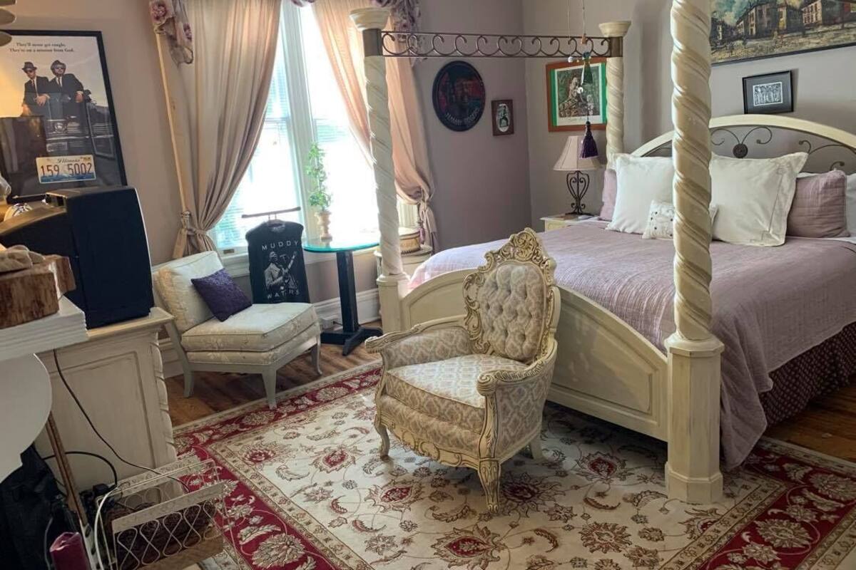 Ein Schlafzimmer mit Himmelbett, einem Sessel, einem Schreibtisch und einem Fenster mit Vorhängen.