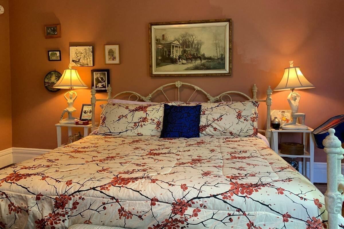 Ein Schlafzimmer mit einem großen Bett, zwei Nachttischlampen, einem gerahmten Gemälde und mehreren kleinen gerahmten Bildern an der Wand.