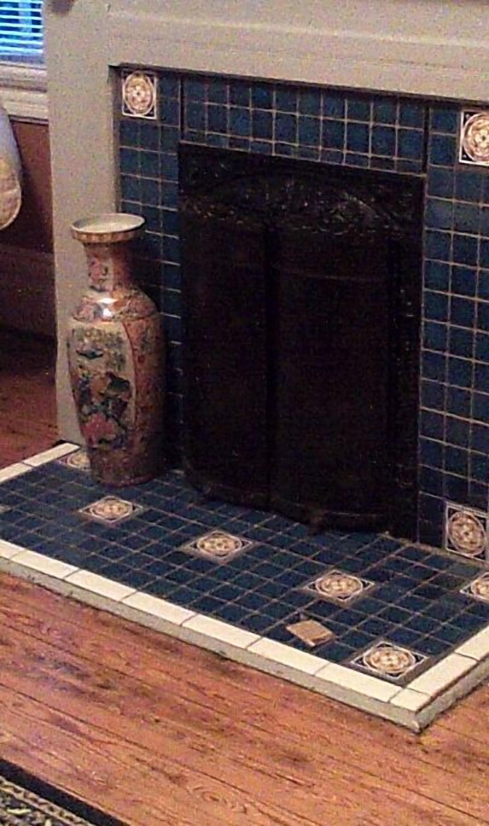 Une cheminée recouverte de carreaux décoratifs, un parquet et un vase placé à gauche.