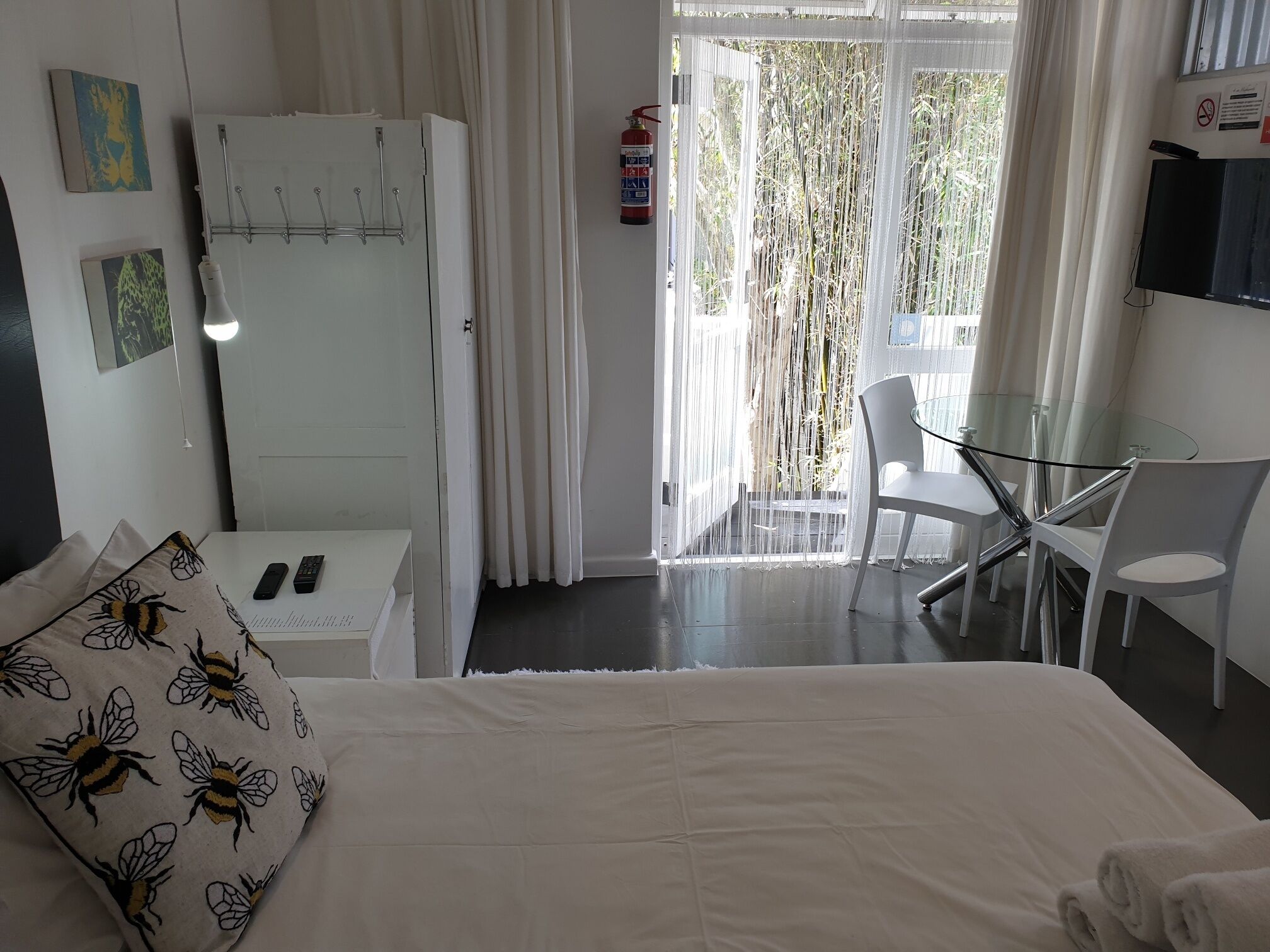 A hotel room with a bed, a desk with a chair, a glass table, a refrigerator, and a window with curtains.