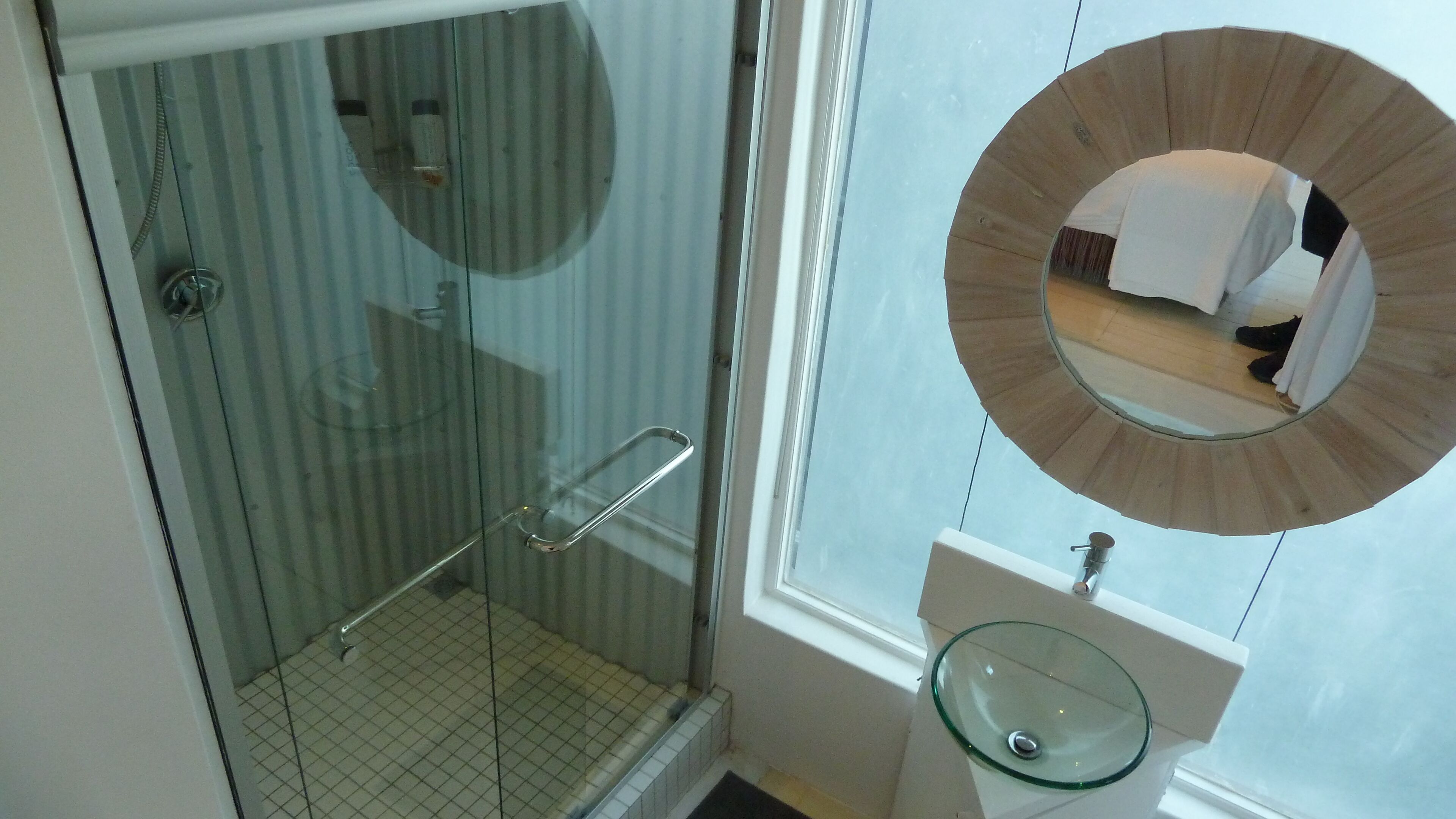 A bathroom with a glass shower enclosure, a glass sink, and a round mirror.