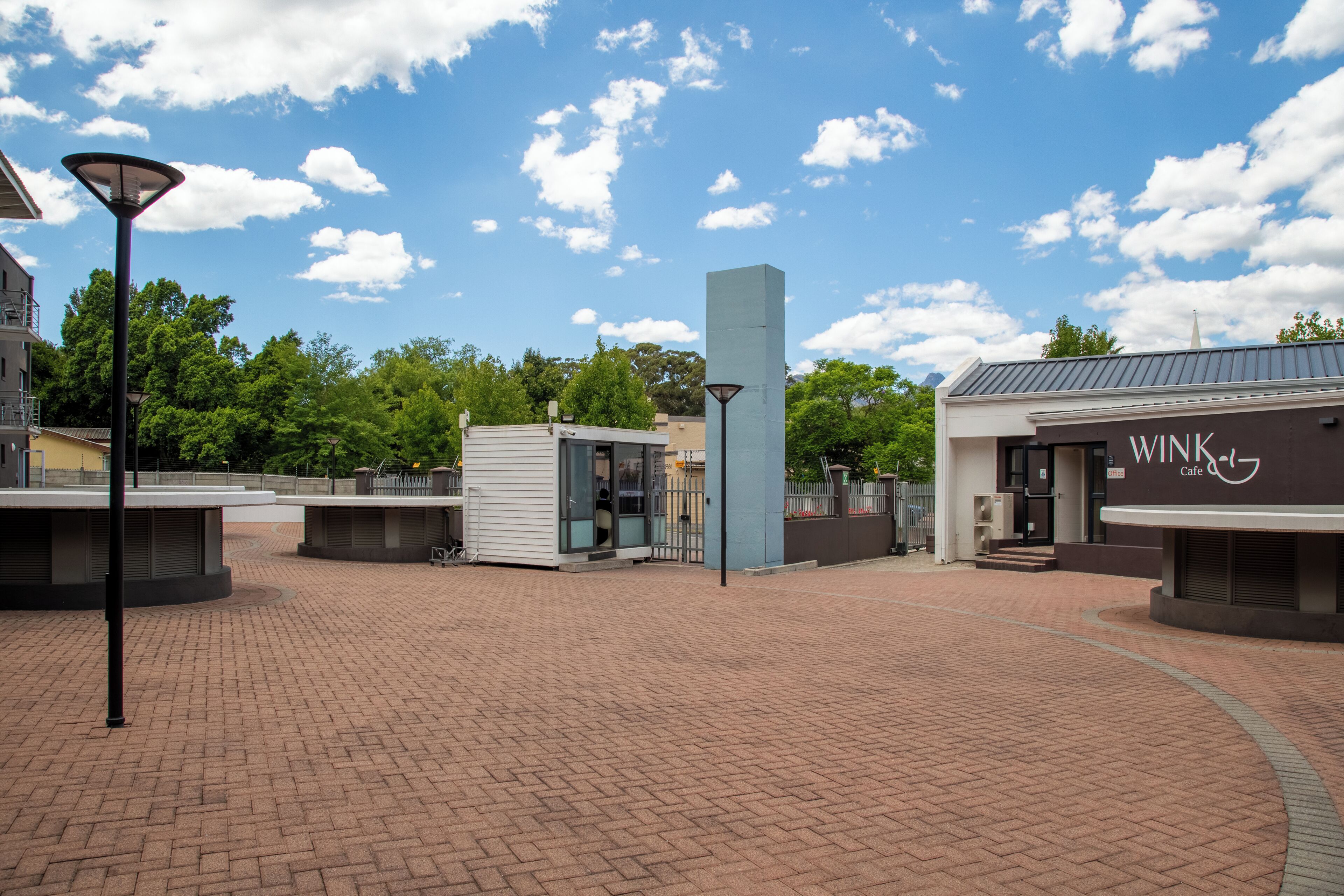 Une place publique avec un bâtiment moderne portant l’inscription « WINK Café », un lampadaire et une zone pavée.