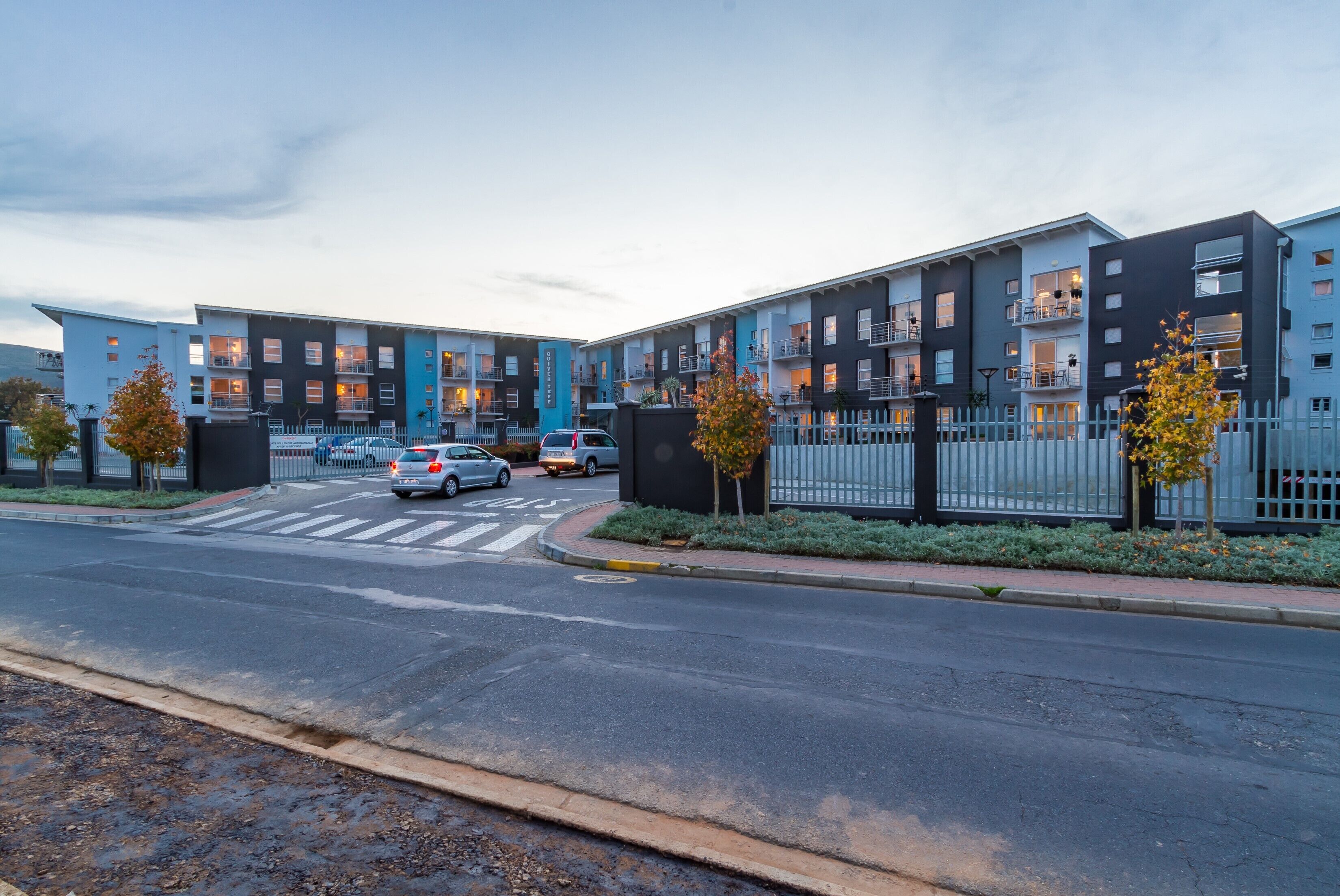 A modern multi-story residential building with balconies, a gated entrance, and a crosswalk.