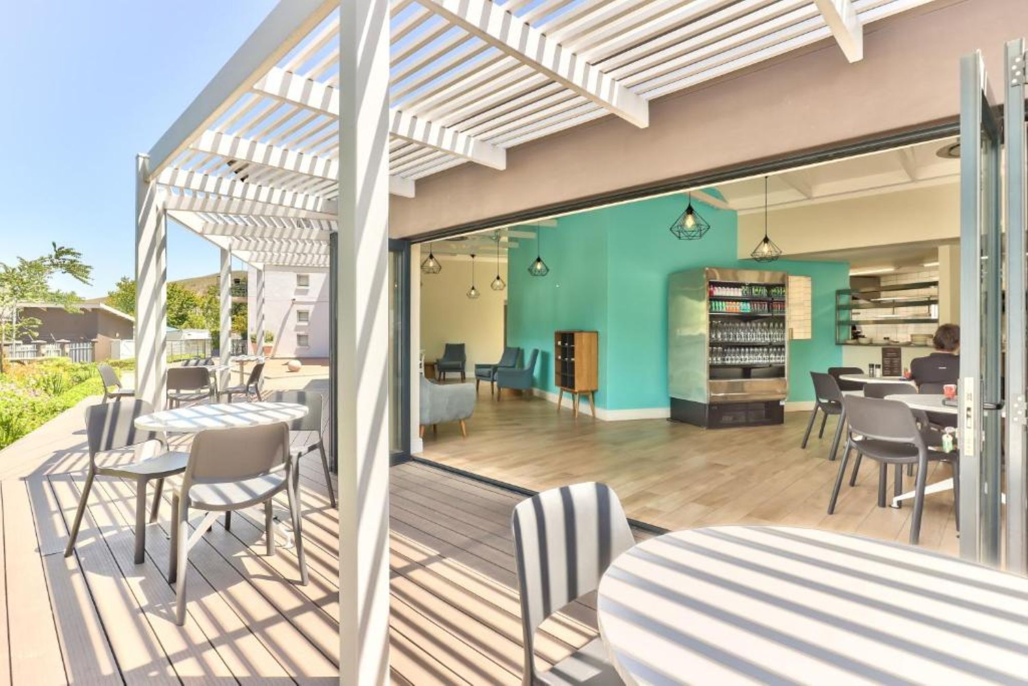 A modern outdoor seating area with tables and chairs, a bar area with a teal wall, and a view of the interior.