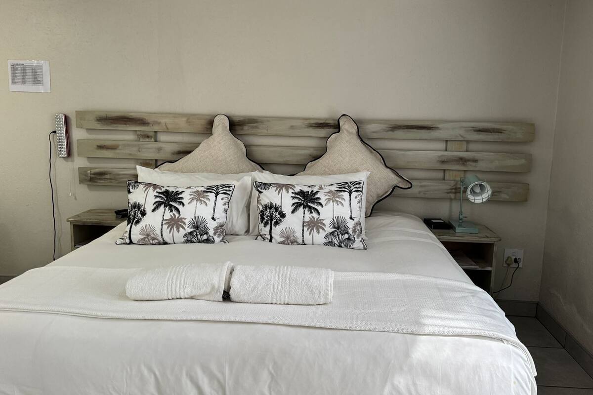 Una cama bien tendida con una colcha blanca y dos almohadas estampadas con motivos tropicales.