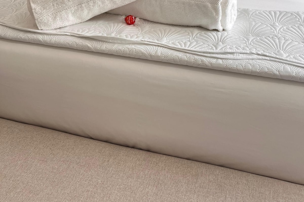 Una cama bien hecha con sábanas blancas y almohadas decorativas, una almohada blanca mullida y un pequeño objeto rojo sobre la cama.