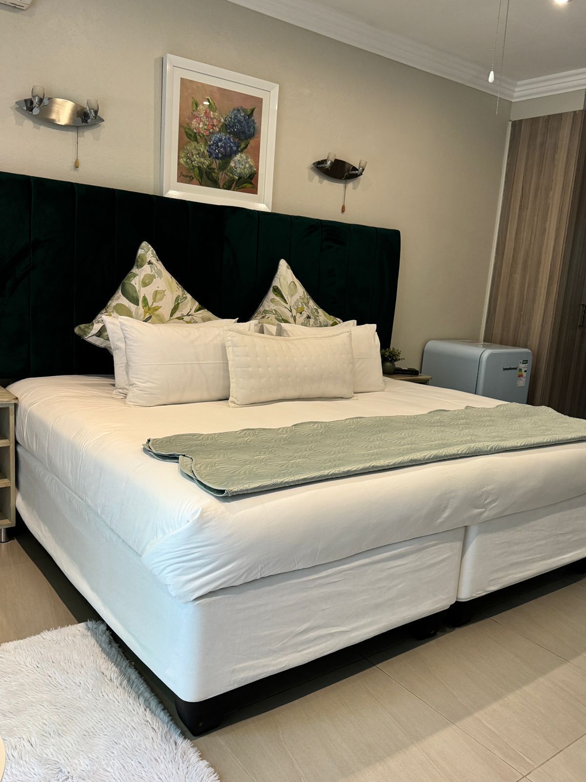 A neatly made bed with white linens and a green throw blanket, a dark headboard, and a framed floral painting on the wall.