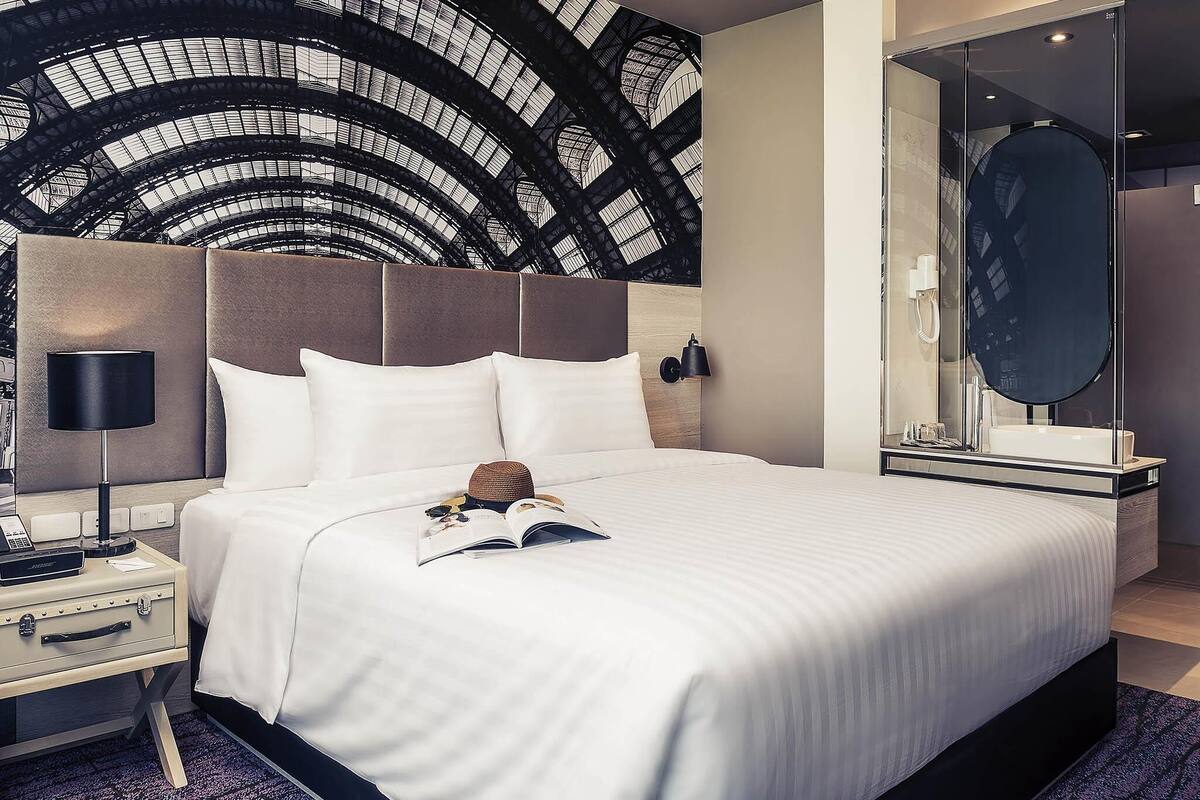 A modern hotel room with a large bed, a nightstand, a lamp, a mirror, and a view of the hotel's architecture.