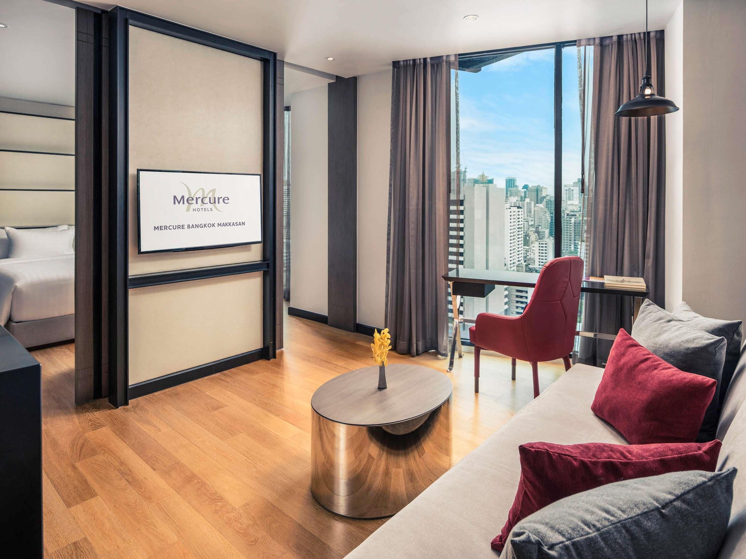 A modern hotel room with a bed, a round coffee table, a red chair, a sofa with red and grey cushions, and a large window with a city view.