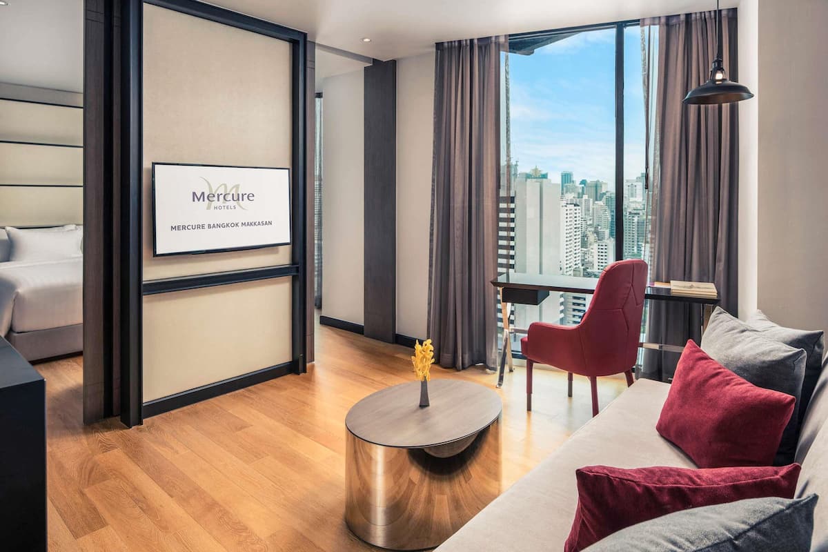 A modern hotel room with a bed, a round coffee table, a red chair, a sofa with red and grey cushions, and a large window with a city view.