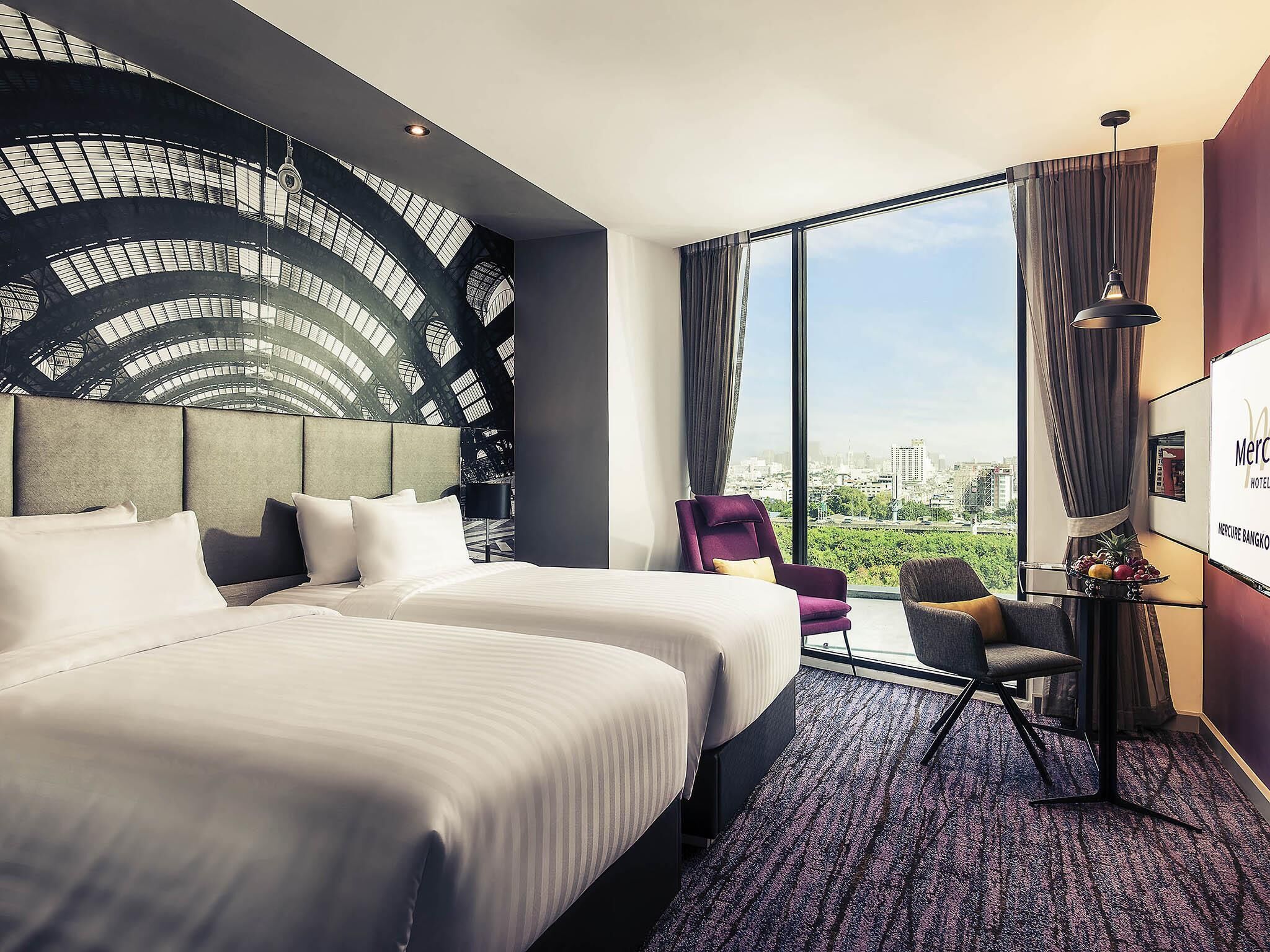 Een hotelkamer met twee bedden, een groot raam met uitzicht op de stad, een modern hoofdeinde en een zithoek met een stoel en een tafeltje.