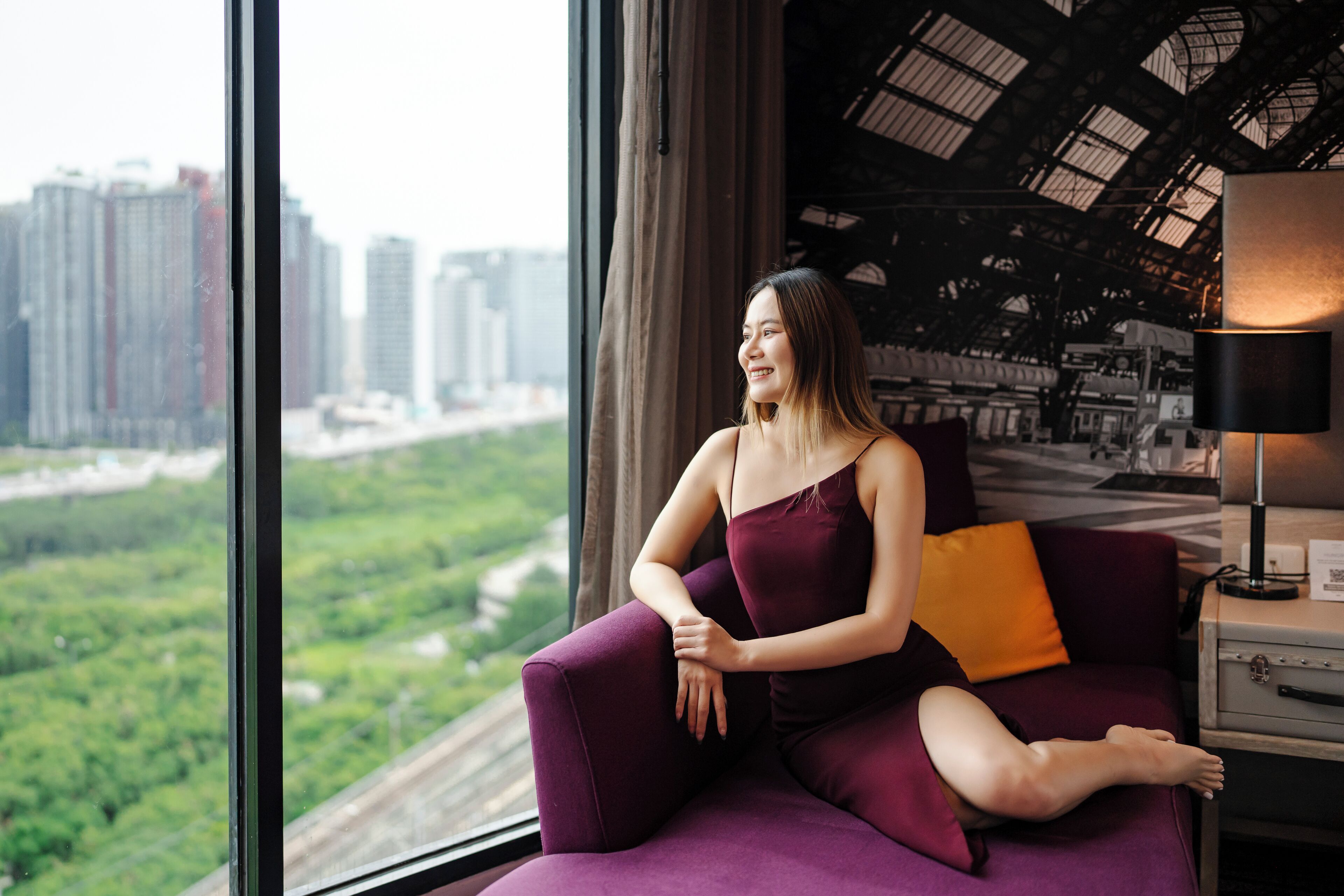 A woman sitting on a purple sofa by a window with a city view.