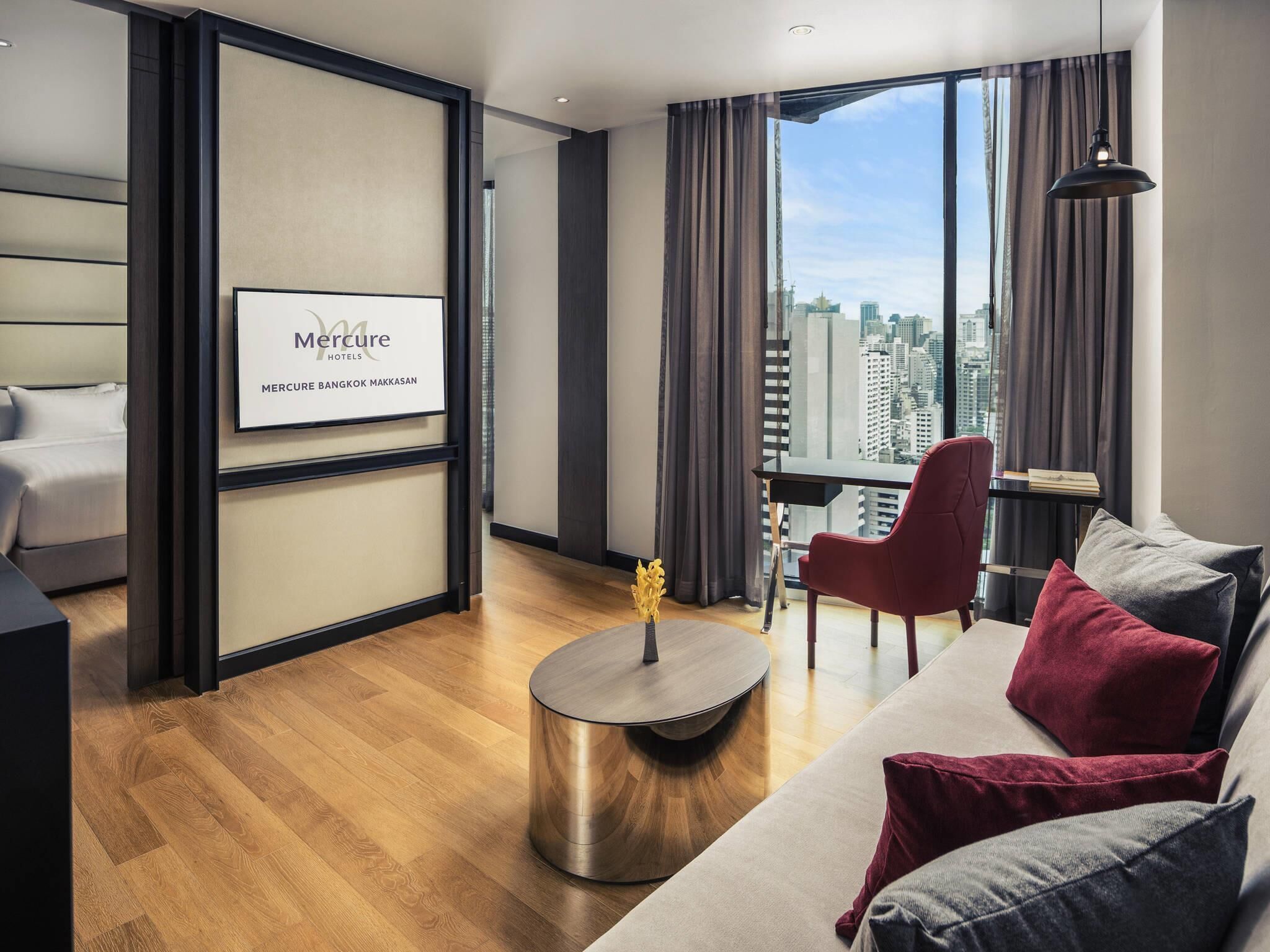 Een moderne hotelkamer met een bed, een bank, een ronde salontafel en een bureau met een stoel. Er is een groot raam met uitzicht op de stad.