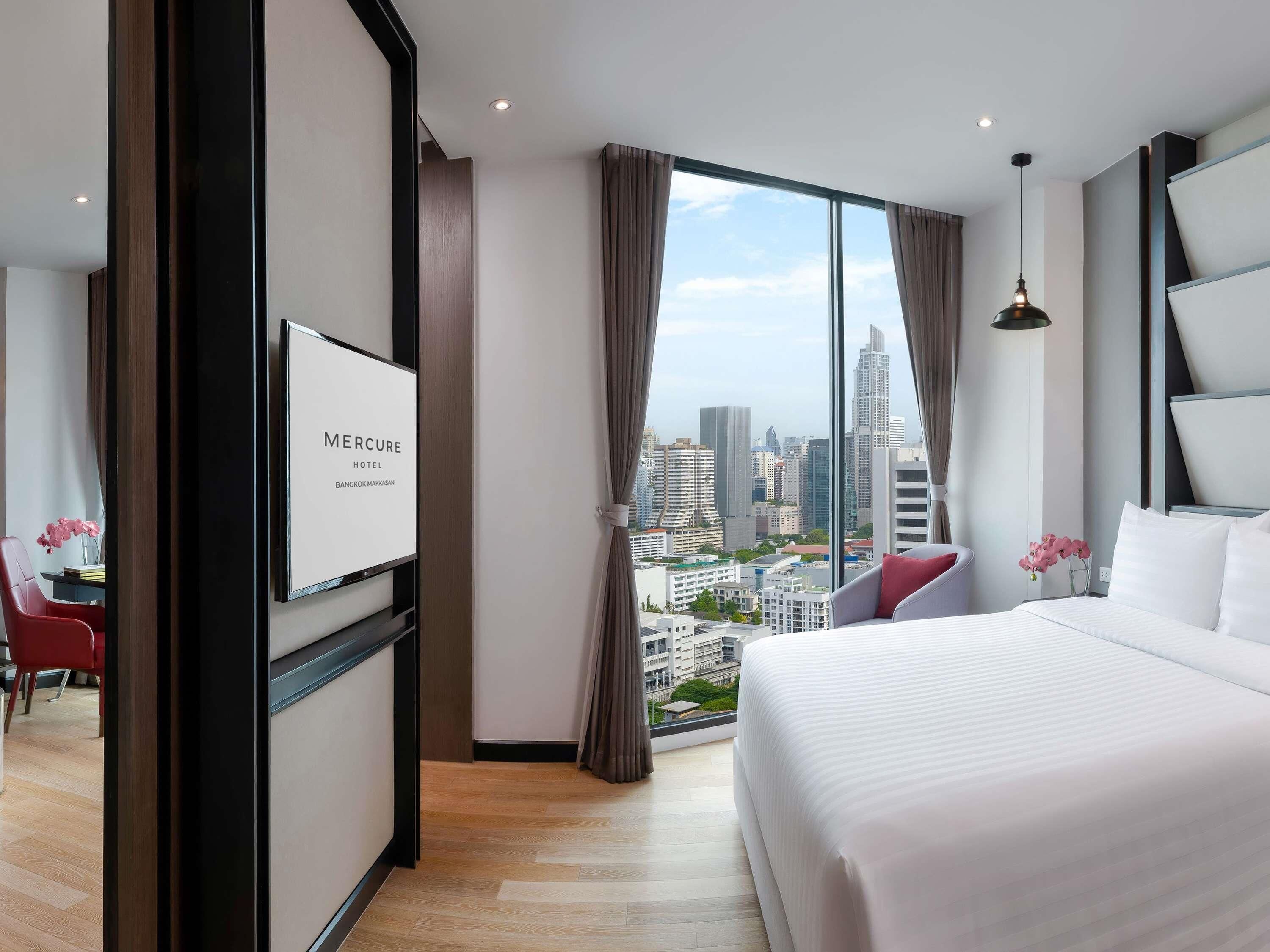 A hotel room with a large bed, a city view, a desk with a chair, and a TV on the wall.