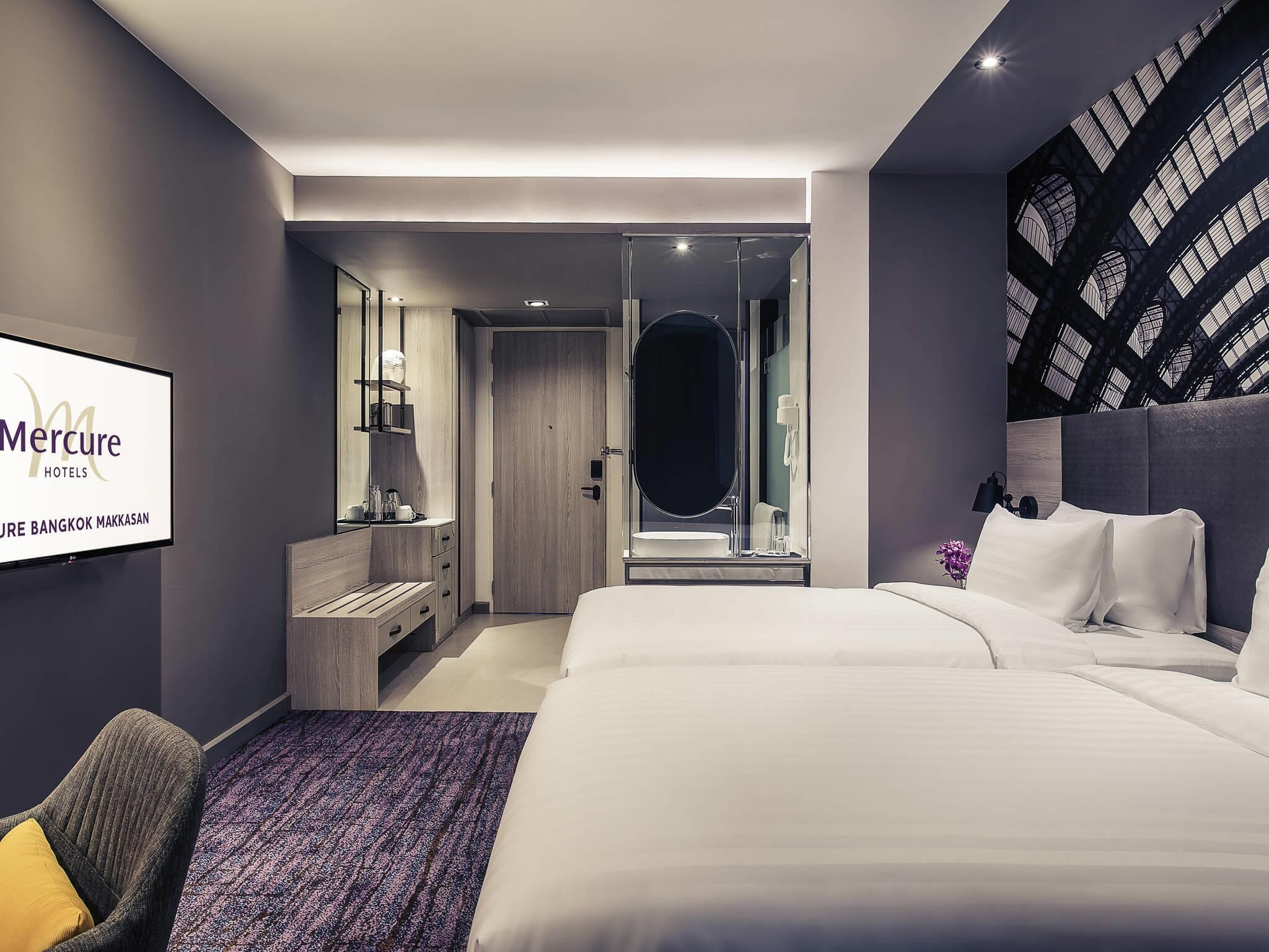 Een moderne hotelkamer met een groot bed, een zithoek, een kaptafel en een televisie waarop de naam van het hotel staat.