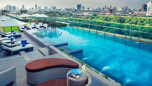 A rooftop infinity pool with lounge chairs and umbrellas, surrounded by greenery and a city skyline in the background. - Mercure Bangkok Makkasan (Bangkok)