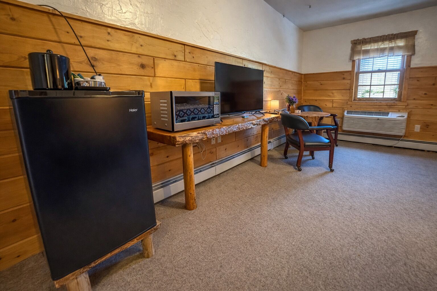 A room with a wooden wall, a refrigerator, a microwave, a television, a table, and a chair.