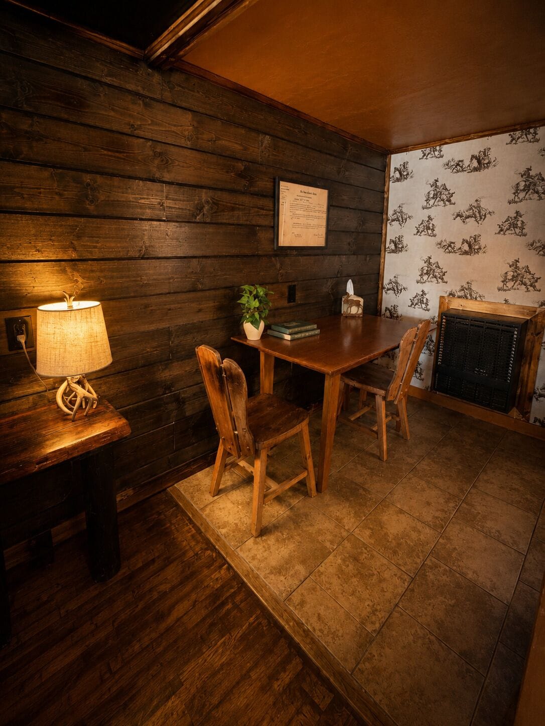A wooden interior with a table and chairs, a lamp, a plant, and a wall with a framed certificate.