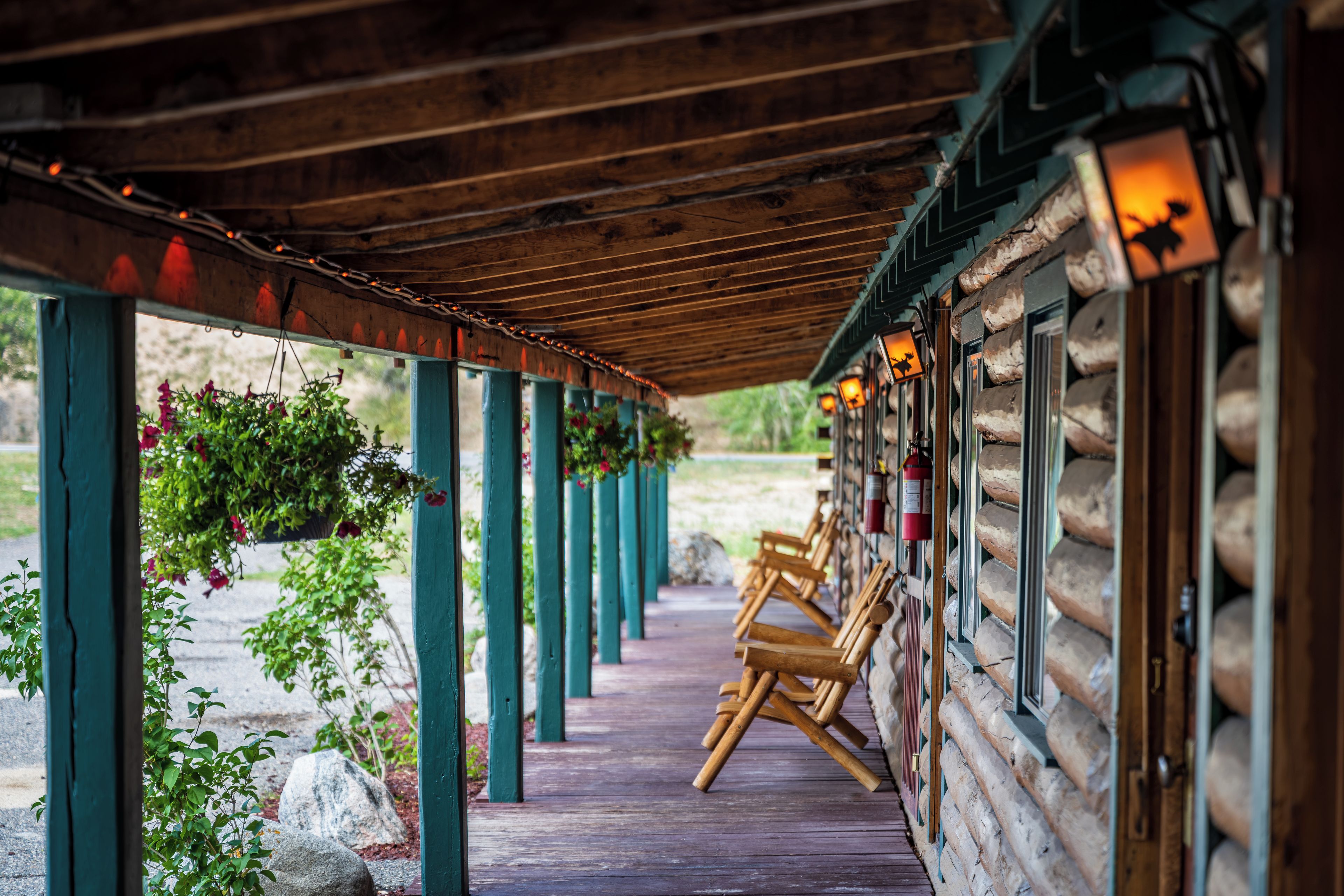 A covered wooden deck with hanging flower baskets, wooden chairs, and a stone wall with windows.