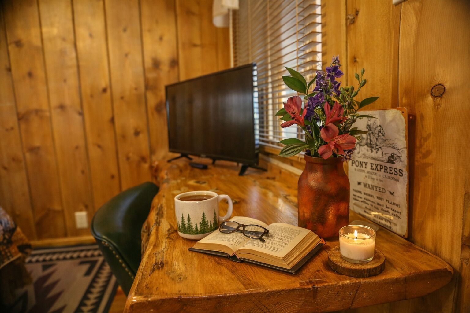 A wooden table with a mug, an open book, a pair of glasses, a candle, and a vase of flowers.