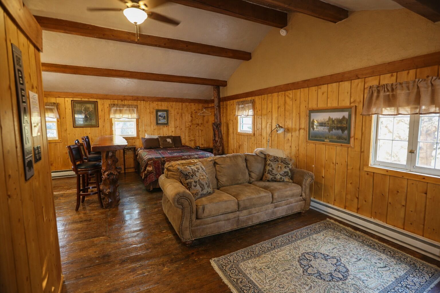 A cozy room with wooden walls and a ceiling fan, a bed with a quilt, a sofa, and a dining table with chairs.