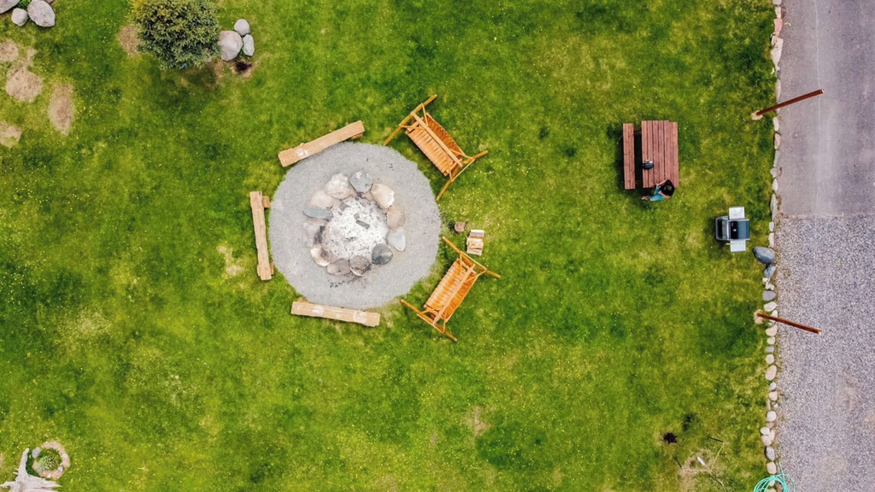 Vista aérea de un espacio de estar circular al aire libre con un brasero, rodeado de bancos de madera, cerca de un camino pavimentado y una estructura de madera.