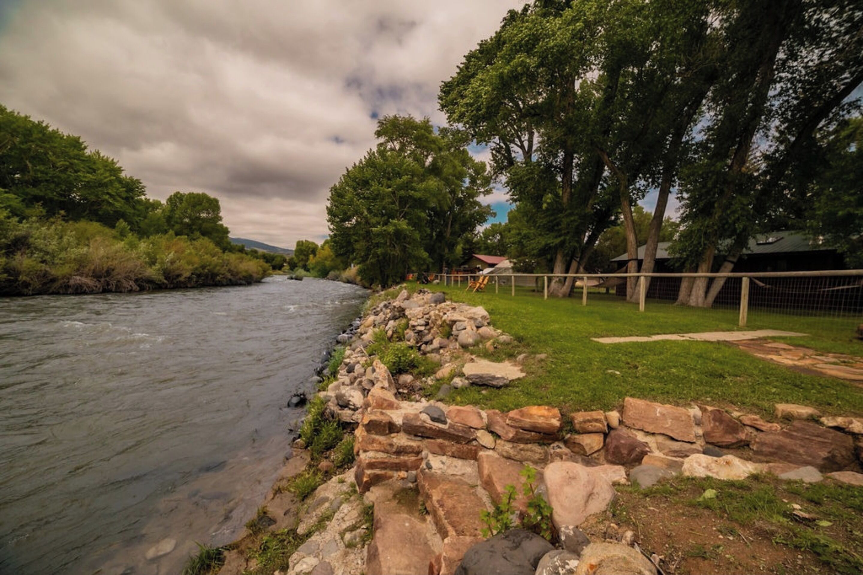 A riverbank with a stone wall, grassy area, and trees.