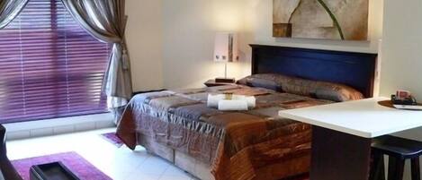 A bedroom with a bed, a desk, a chair, a window with curtains, and a painting on the wall.