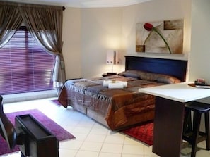 A bedroom with a bed, a desk, a chair, a window with curtains, and a painting on the wall.