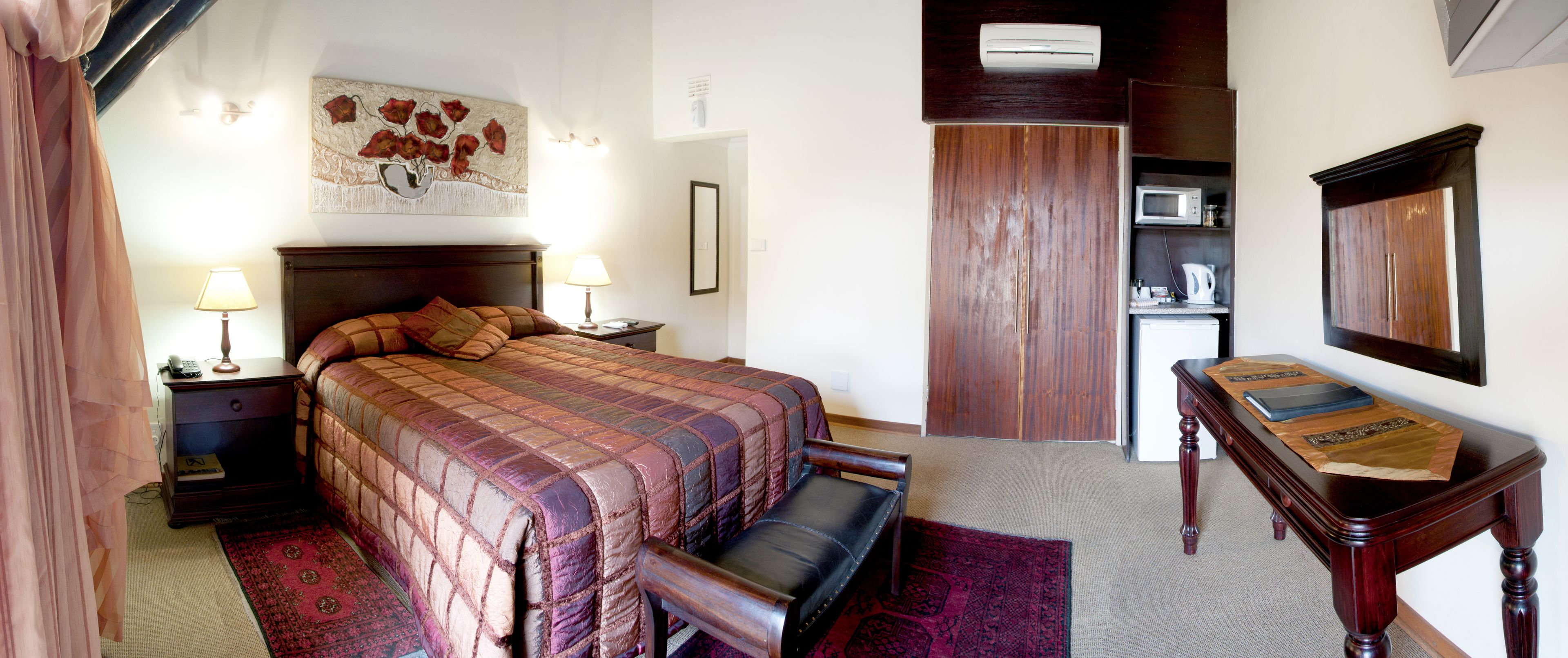 A hotel room with a large bed, bedside lamps, a wooden nightstand, a desk with a chair, a microwave, and a wardrobe.