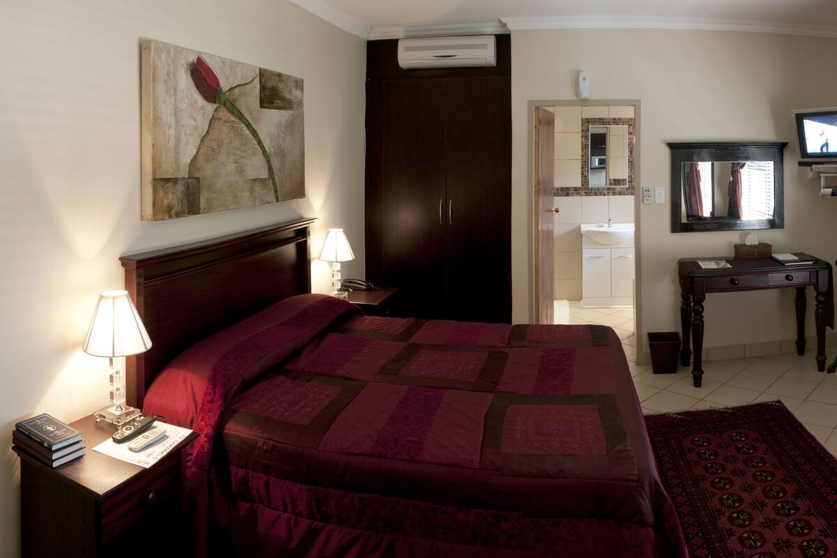 A hotel room with a large bed, a nightstand with a lamp, a desk with a chair, a television, and a bathroom visible through an open door.