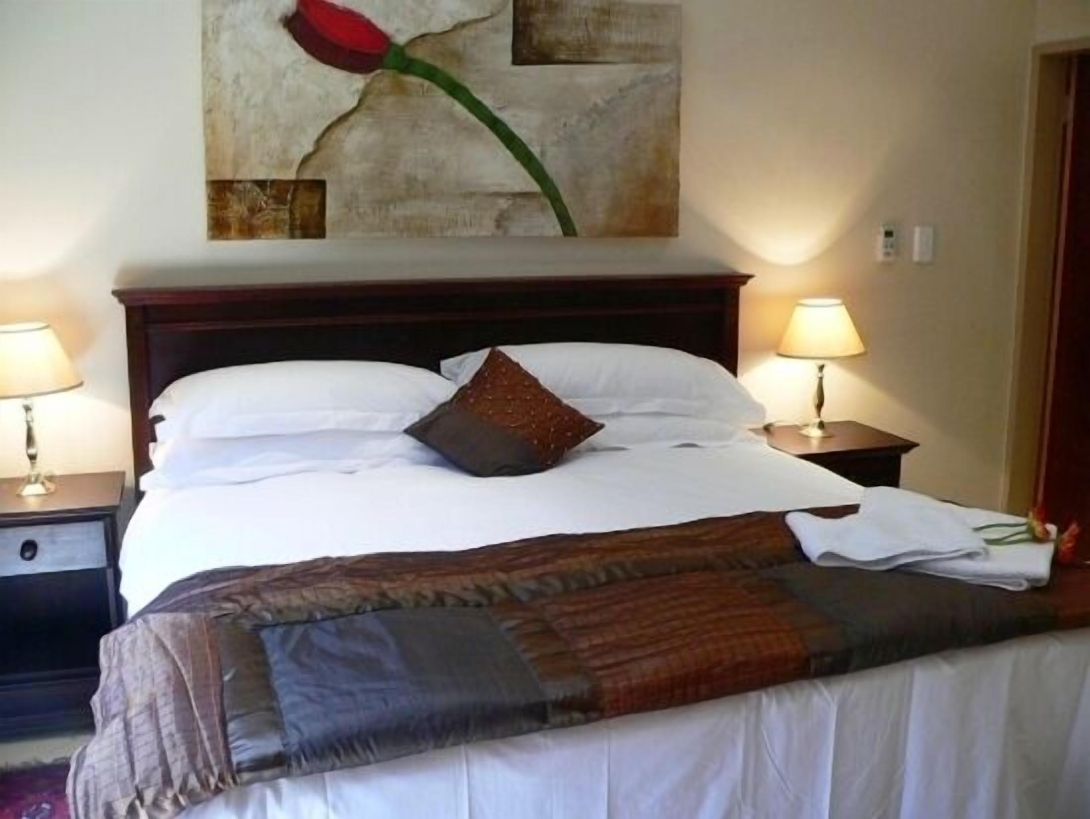 A neatly made bed with white linens and a brown throw blanket, two bedside lamps, and a wall-mounted painting of a red rose.