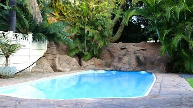 A swimming pool with a stone border, surrounded by lush greenery and a white fence. - Rustenburg Boutique Hotel (Rustenburg)