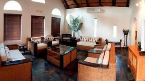 A spacious living area with multiple seating arrangements, including sofas and a coffee table. There are large windows with blinds, a potted plant, and a ceiling fan. - Rustenburg Boutique Hotel (Rustenburg)