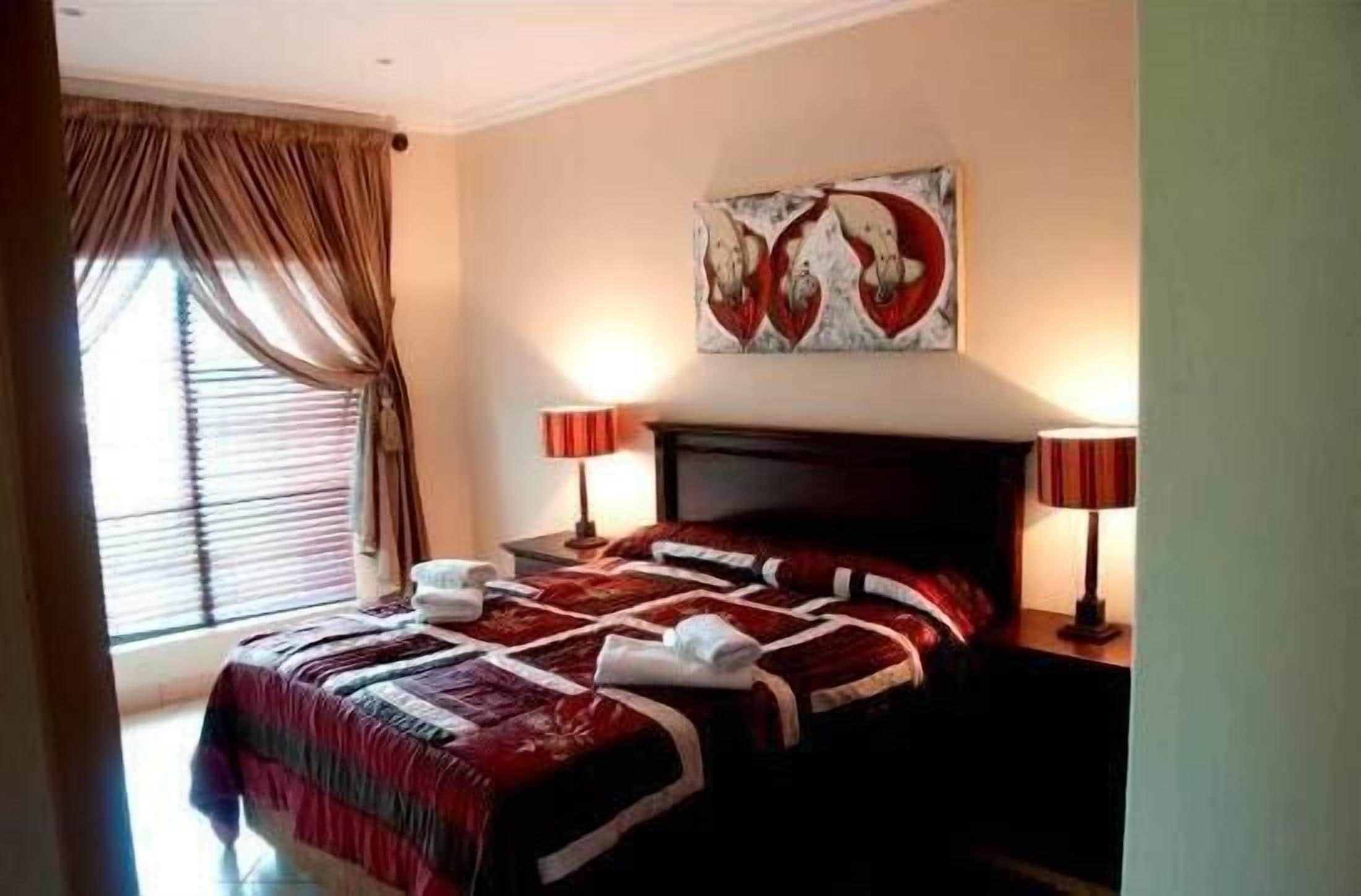 A hotel room with a large bed, two bedside lamps, a painting on the wall, and a window with curtains.
