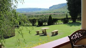 A covered outdoor area with a table and chairs, overlooking a green landscape with hills. - Oranje Gasteplaas Maloti Route (Fouriesburg)