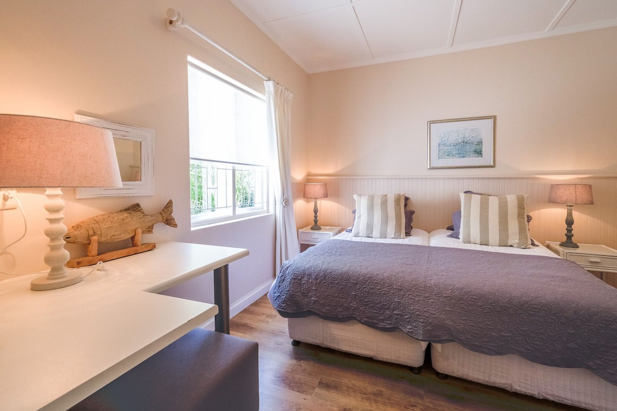 Un dormitorio con cama, mesitas de noche, una lámpara, una ventana con cortinas, un cuadro enmarcado en la pared y una escultura de pez en la mesita de noche.
