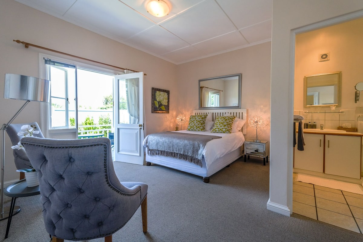 Un dormitorio con cama, una silla, mesita de noche, lámpara, ventana y baño con espejo y mueble.