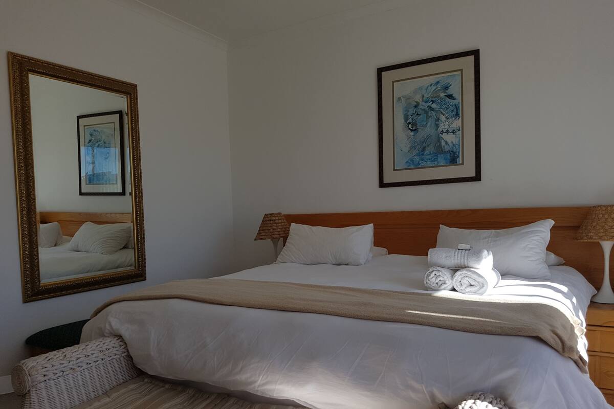 Cama bem arrumada com lençóis brancos, cabeceira de madeira e um espelho grande com moldura dourada.