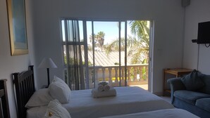 A hotel room with two beds, a balcony with a view of palm trees, a TV on the wall, and a blue sofa. - Melkbosch Guesthouse (Cape Town)