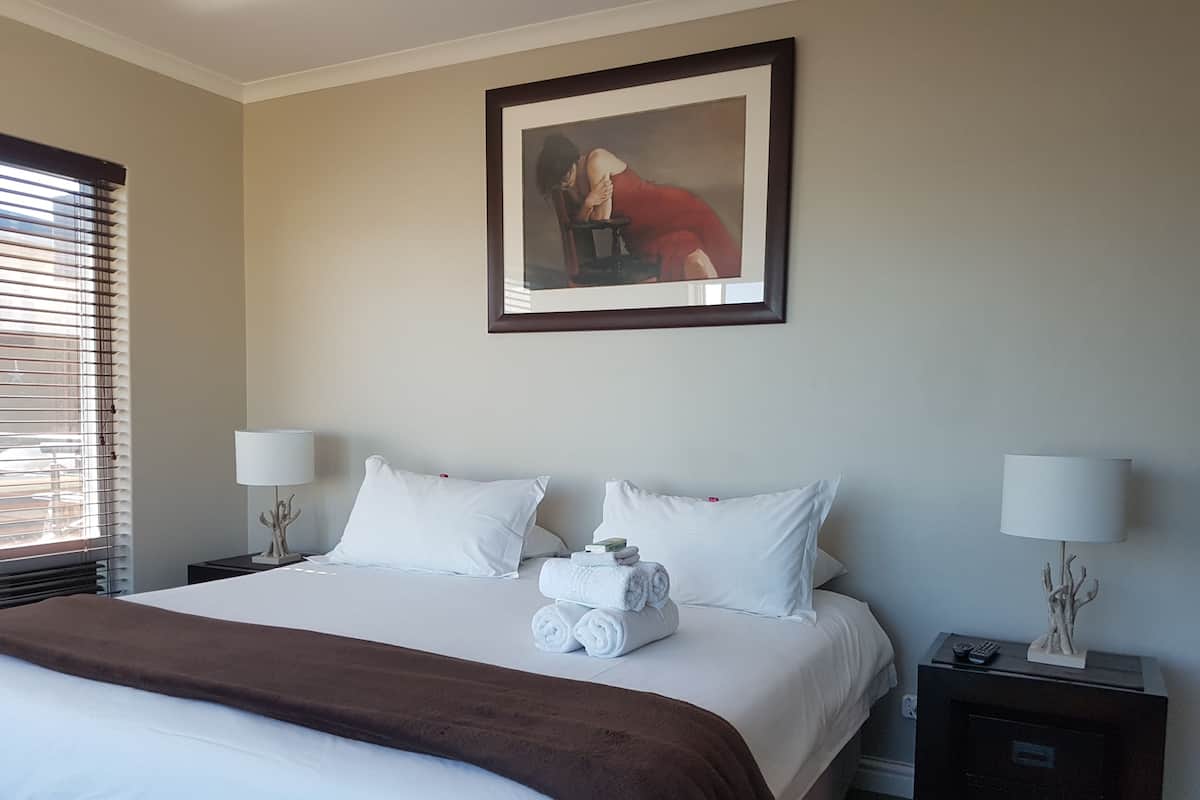 Quarto de hotel com uma cama grande, duas mesas de cabeceira com luminárias, um quadro na parede e uma janela com persianas.