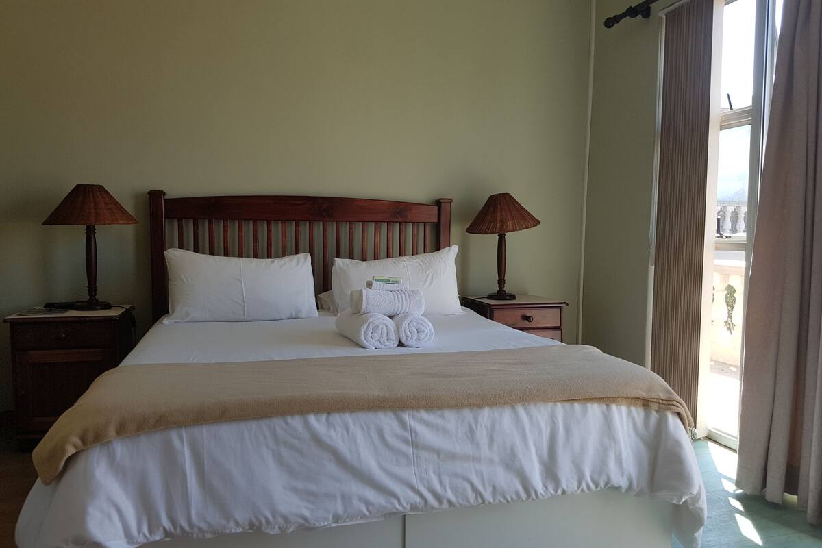 Uma cama bem arrumada com lençóis brancos e um cobertor bege, acompanhada por duas mesinhas de cabeceira de madeira com luminárias, em um quarto com uma grande janela e cortinas.